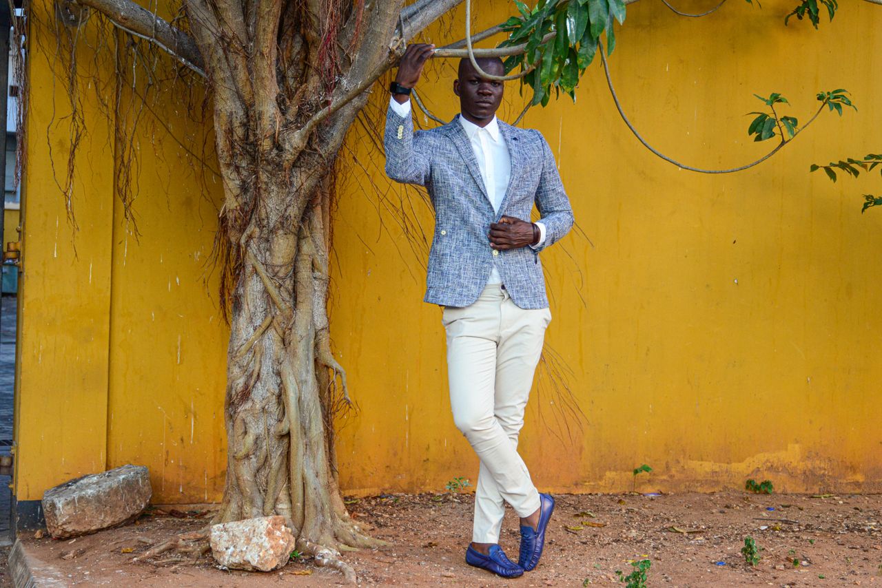 gallery-03 Fashionable man in blazer poses by a tree against a vibrant yellow wall.