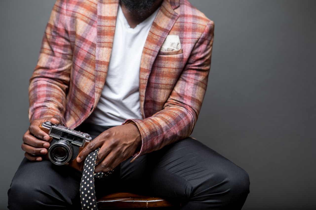 home-img A fashionable man in a plaid blazer holds a vintage camera, seated indoors.