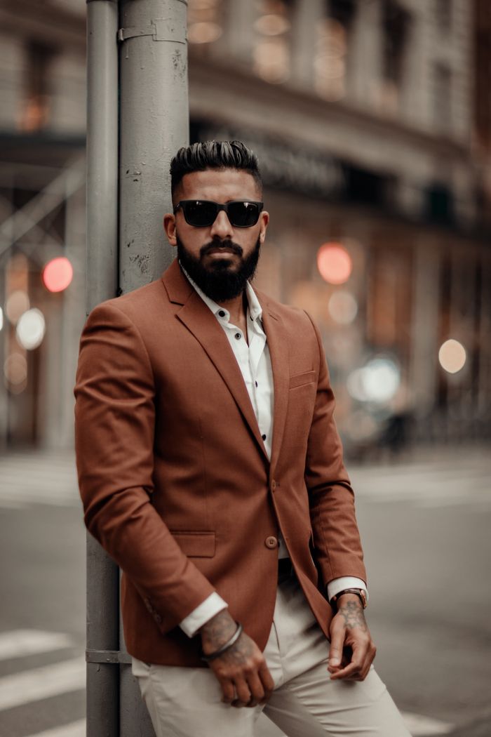 gallery-05 Man with sunglasses posing elegantly in an urban setting wearing a brown blazer.