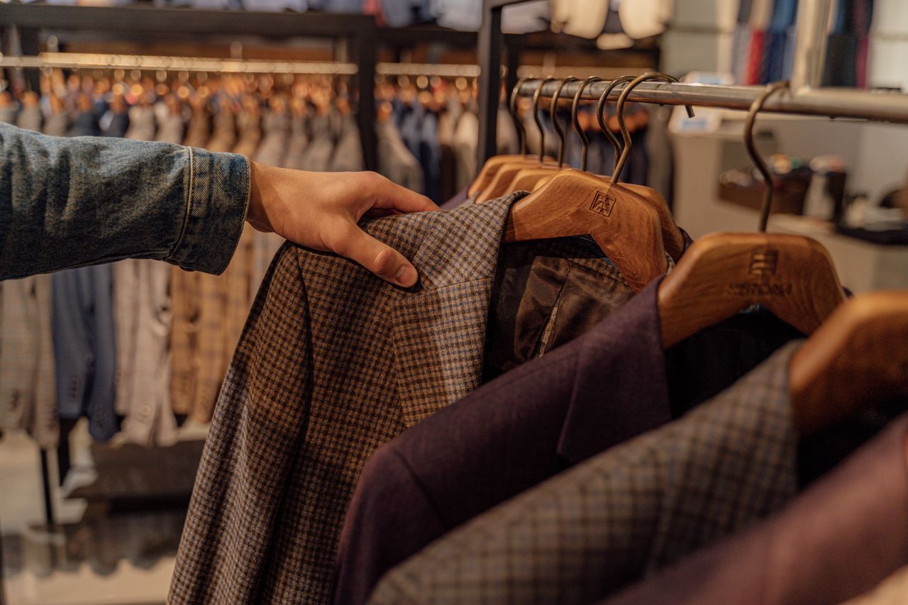 about-img-01 Close-up of a hand selecting men's blazers in a fashion store, highlighting trendy styles.