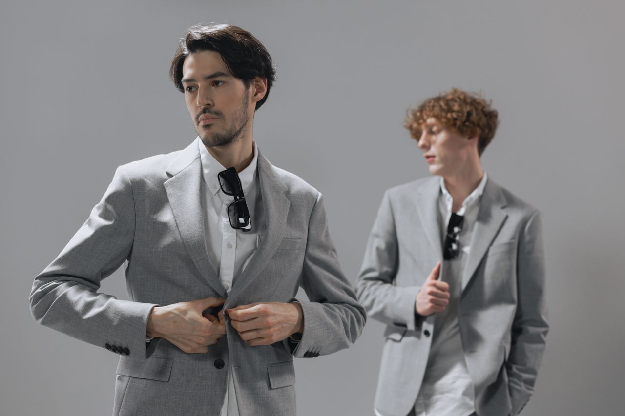 gallery-02 Studio shot of two men in gray suits posing confidently with sunglasses indoors.
